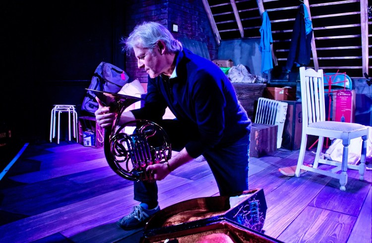 A silver-haired middle-aged man wearing a dark sweater, jeans and running shoes, taking a knee on the wooden floor of an attic, holding a French horn and gazing into the bell. Old furniture, boxes and clothes in the background.