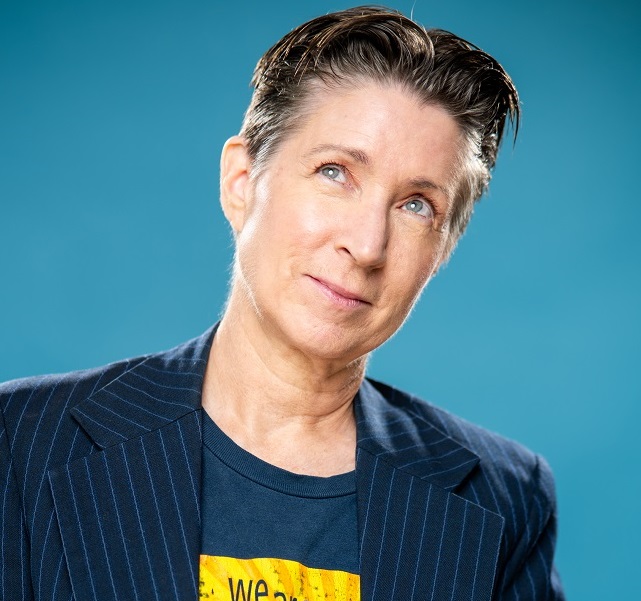 A woman with short brown hair greying at the temples, wearing t-shirt and a navy pinstripe blazer, gazes up to her right with a glint in her eye.